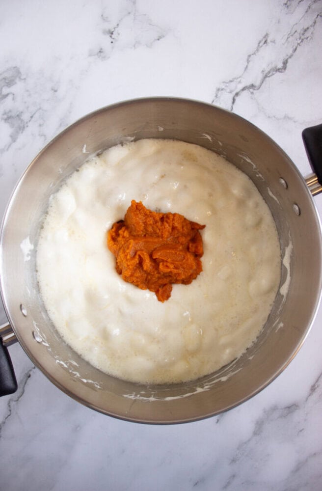A saucepan containing a creamy white mixture with a dollop of orange pumpkin puree in the center, placed on a marble countertop.