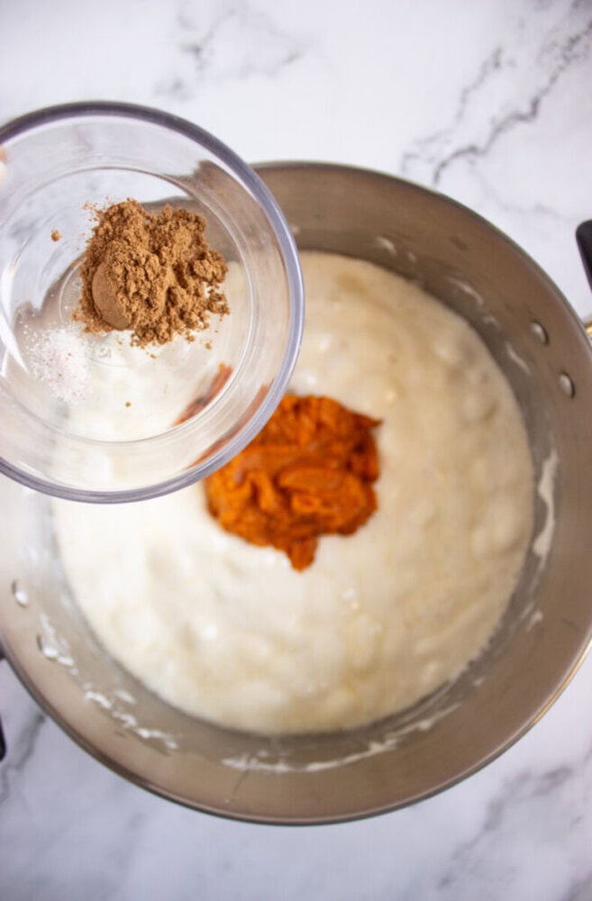 A mixing bowl with batter and pumpkin puree, with a small bowl of spices and salt being held above, ready to add.