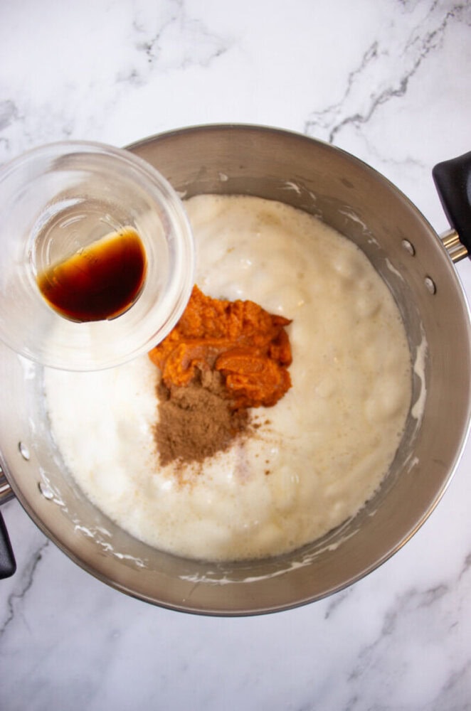 A metal mixing bowl with creamy batter, pumpkin puree, and spices, while a hand pours vanilla extract from a small glass cup into the mixture.