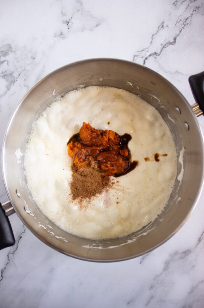 A stainless steel pot contains a creamy white mixture, with pumpkin puree, brown spices, and dark liquid added on top, on a marble countertop.