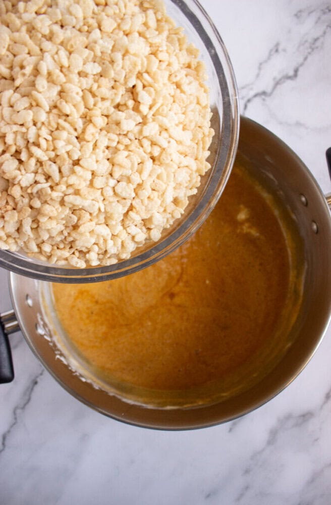 A glass bowl of rice cereal being poured into a saucepan containing melted mixture on a marble countertop.