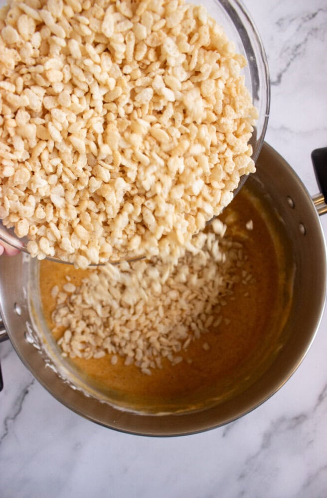 A bowl of puffed rice cereal is being poured into a pot containing a melted, light brown mixture on a marble countertop.