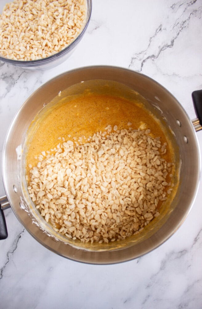 A metal pot contains a yellow mixture with Rice Krispies cereal partially stirred in; a bowl of plain Rice Krispies sits nearby on a marble surface.
