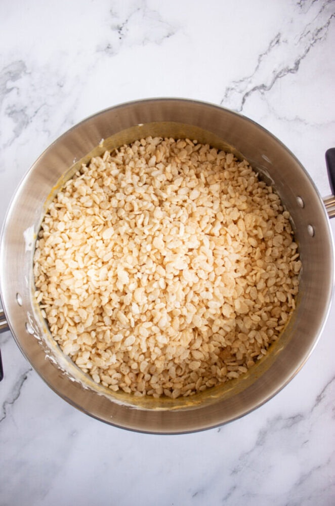 A metal pot filled with rice crispy cereal mixed with melted marshmallows, placed on a marble countertop.
