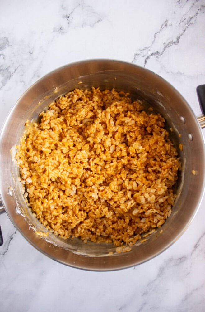 A large metal pot filled with mixed rice cereal coated in a brown, sticky mixture sits on a marble countertop.
