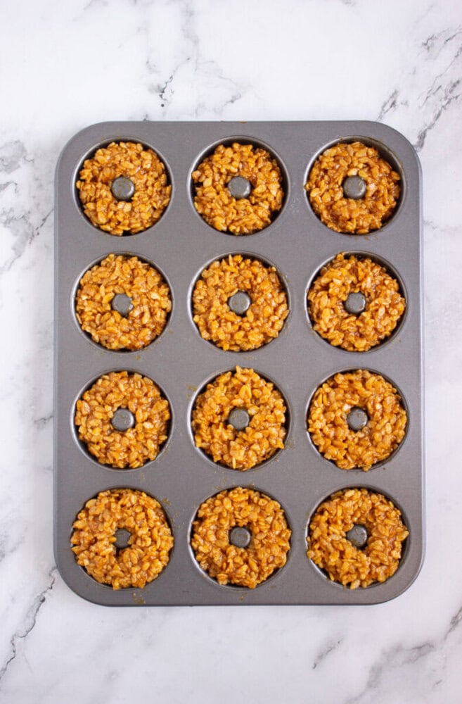 A donut-shaped baking pan filled with an even layer of brown, crispy rice treat mixture in each mold, placed on a white marble surface.
