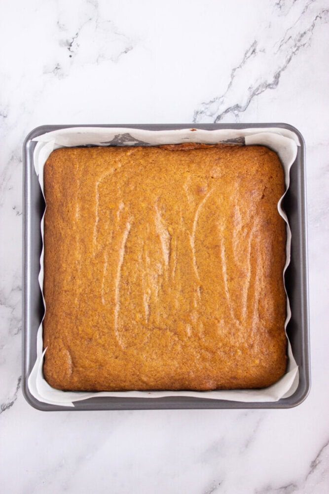 A square pan lined with parchment paper holds a freshly baked, golden-brown sheet cake on a marble surface.