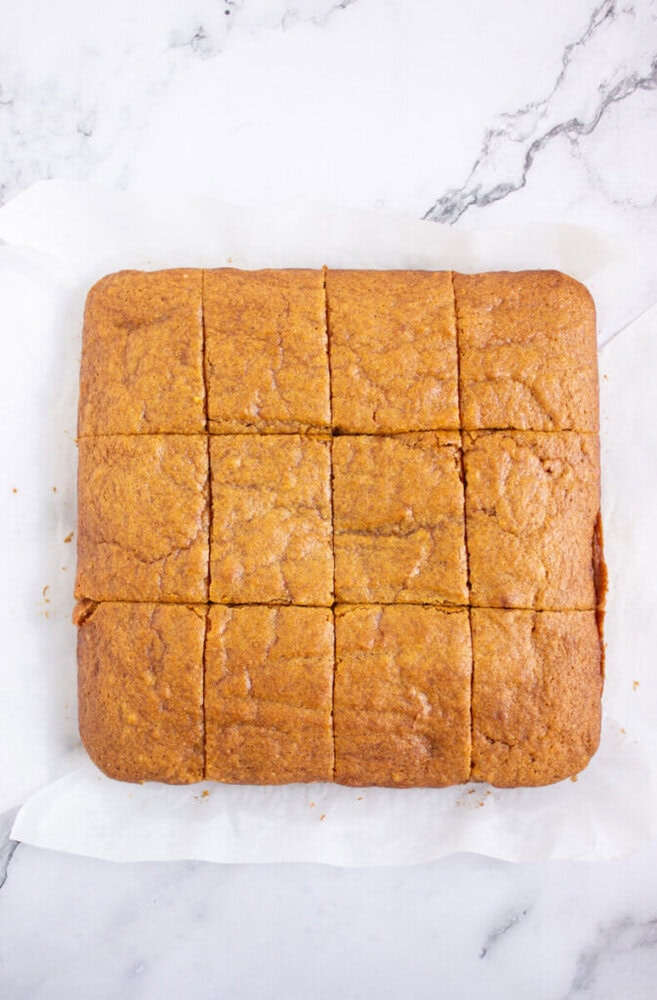 A square, golden-brown cake cut into twelve even pieces sits on white parchment paper on a marble surface.