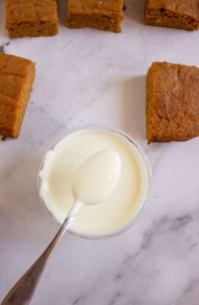 A bowl of white icing with a spoon rests next to several square pieces of cake on a white marble surface.