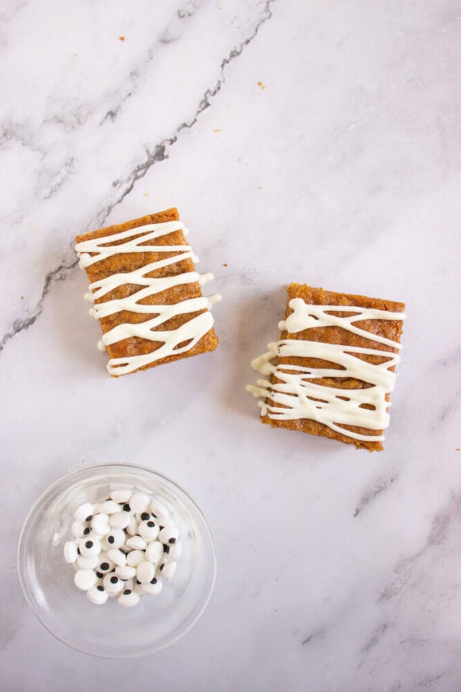 Two rectangular cookie bars with white icing drizzle on a marble surface, next to a small glass bowl containing candy googly eyes.