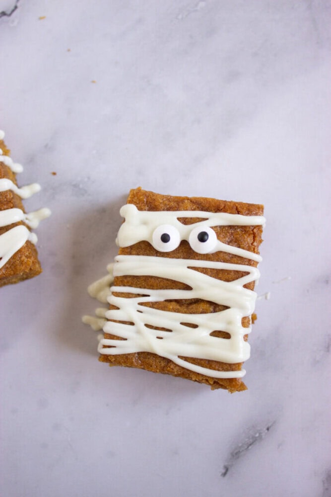 A rectangular bar topped with white icing in a zigzag pattern and two candy eyes, resembling a mummy, on a light marble surface.