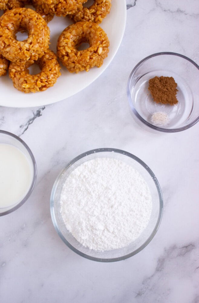 Bowls of powdered sugar, milk, spices, and a plate of ring-shaped treats are arranged on a white marble surface.