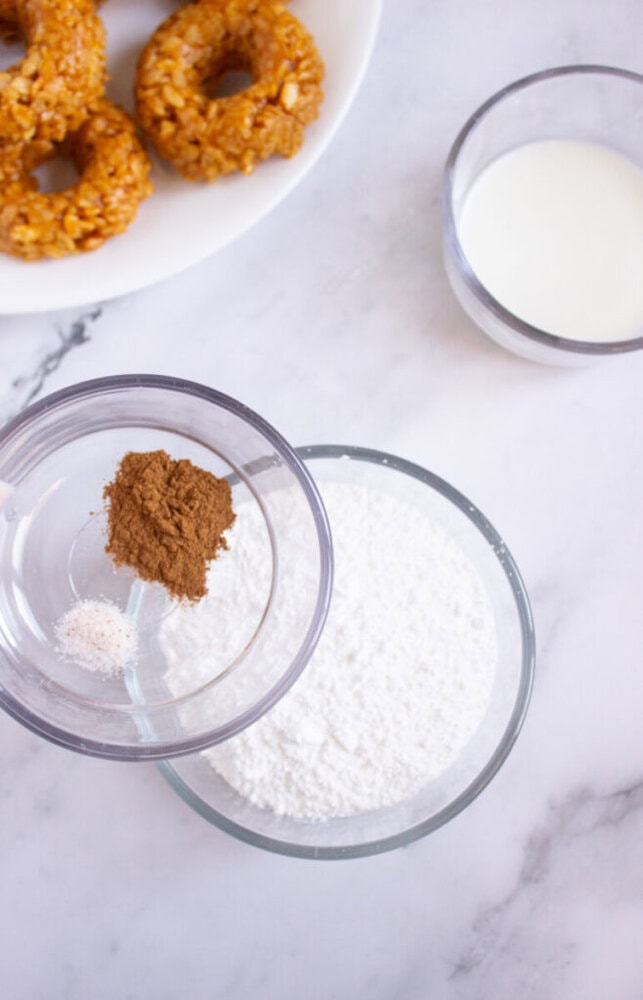 Three glass bowls containing cinnamon, flour, and milk are arranged on a marble surface, with a plate of donuts partially visible in the upper left corner.