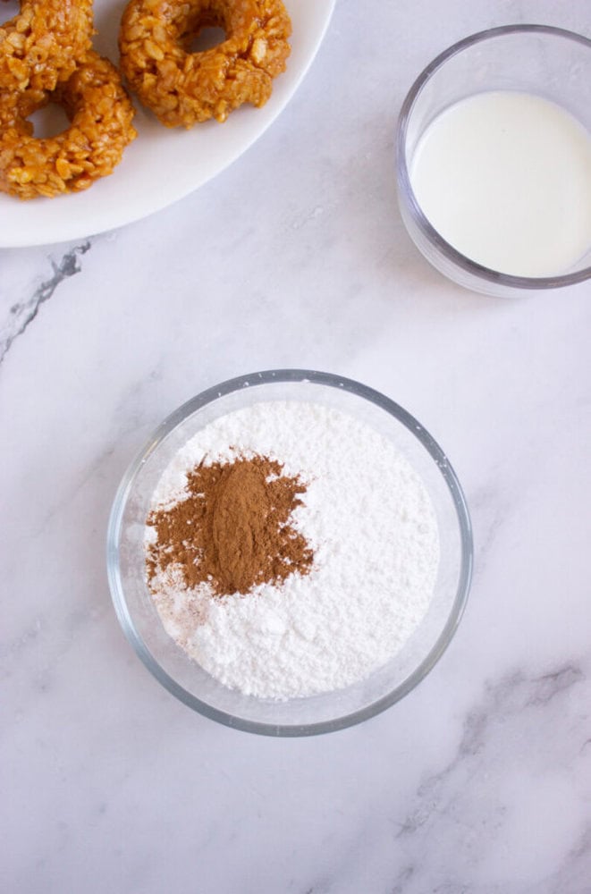 A bowl with powdered sugar and cinnamon, a glass of milk, and a plate with donuts on a white marble surface.