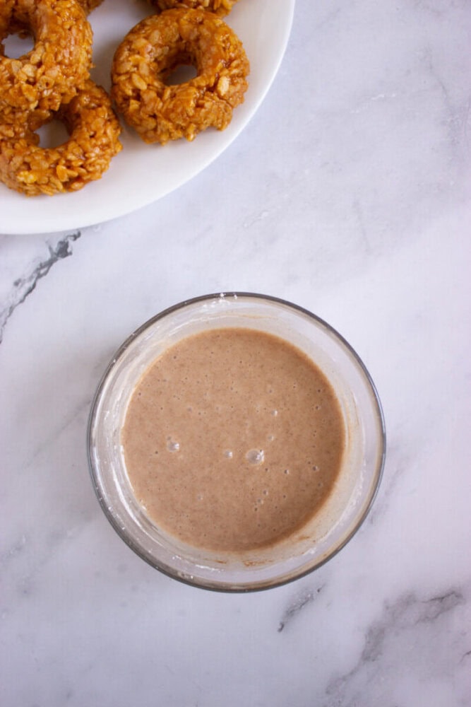 A glass bowl of light brown glaze on a marble surface with several oat donuts on a white plate in the corner.