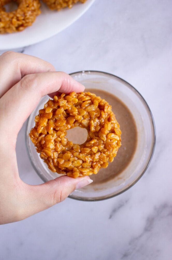 A hand holds a round rice krispie treat over a glass of milk, with another treat on a plate in the background.