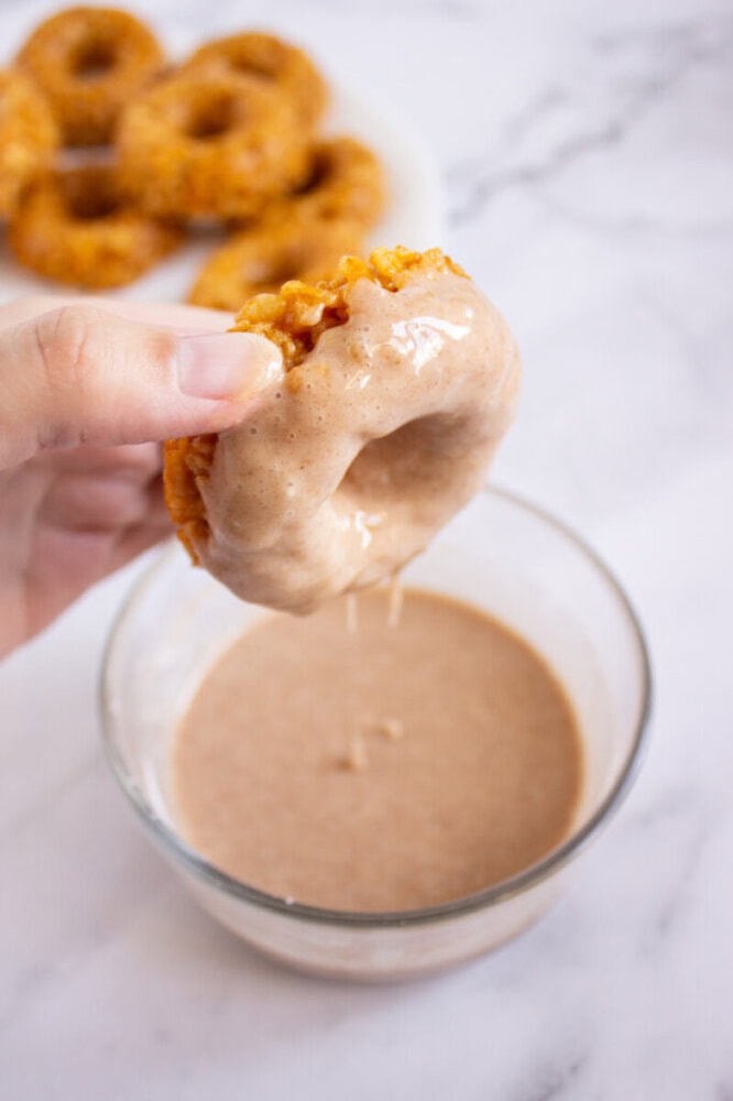 A hand dips a donut into a bowl of light brown glaze, with more donuts on a plate in the background.