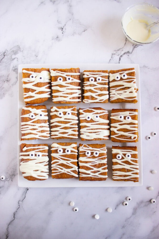 A plate of twelve rectangular brownies decorated with white icing and candy eyes to resemble mummies, next to a bowl of icing and scattered candy eyes on a marble surface.