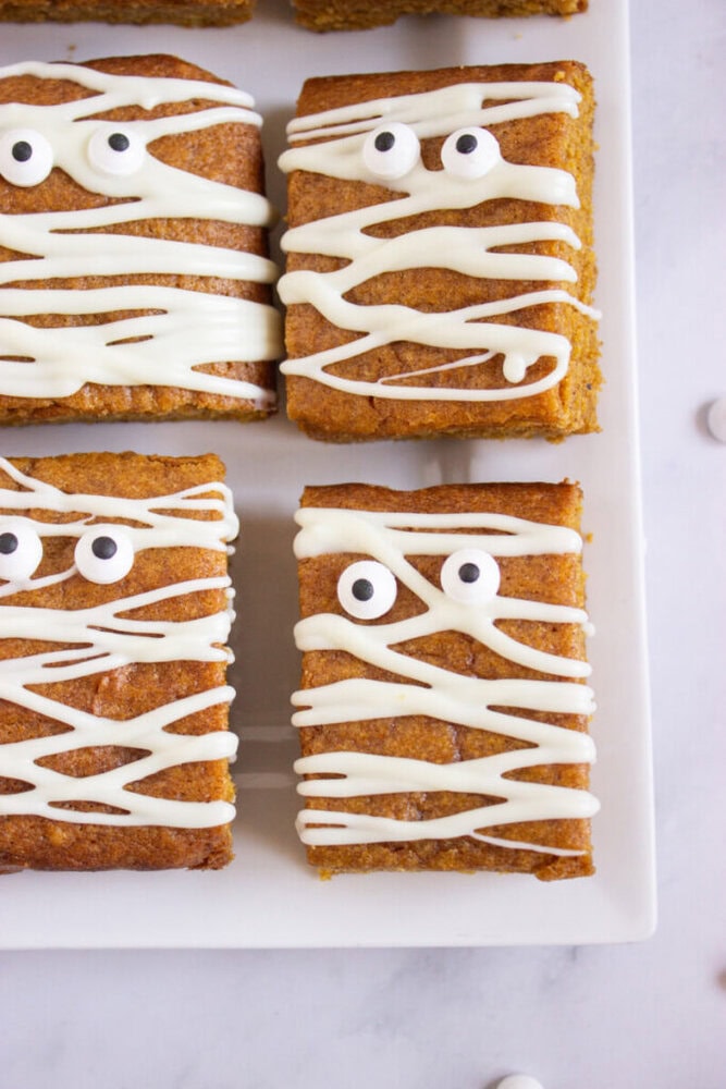 Four rectangular pumpkin bars with white icing drizzled in a mummy-like pattern and candy eyes on a white plate.