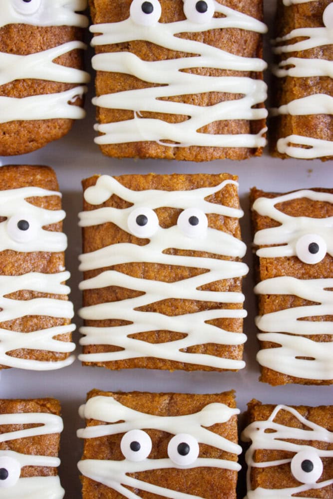 Square baked treats decorated with zigzagged white icing and candy eyes to resemble mummies, arranged in rows on a white surface.