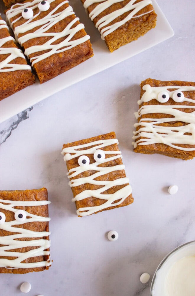 Rectangular baked treats decorated with white icing in a zigzag pattern and candy eyes, arranged on a marble surface and a white plate.