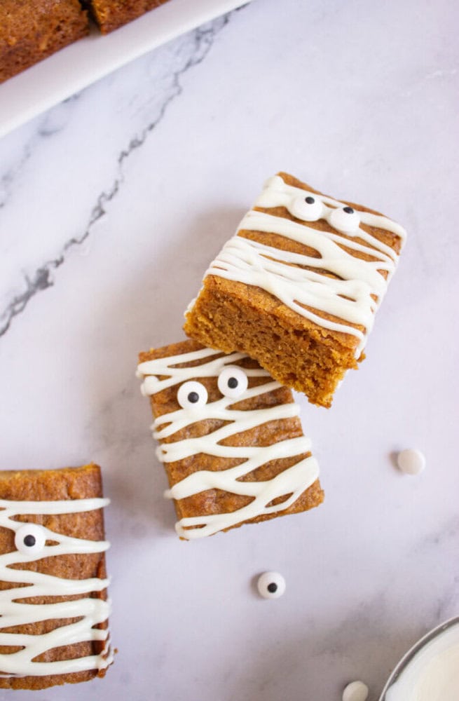Two rectangular pumpkin bars decorated with white icing in a mummy pattern and candy eyes, placed on a light marble surface.