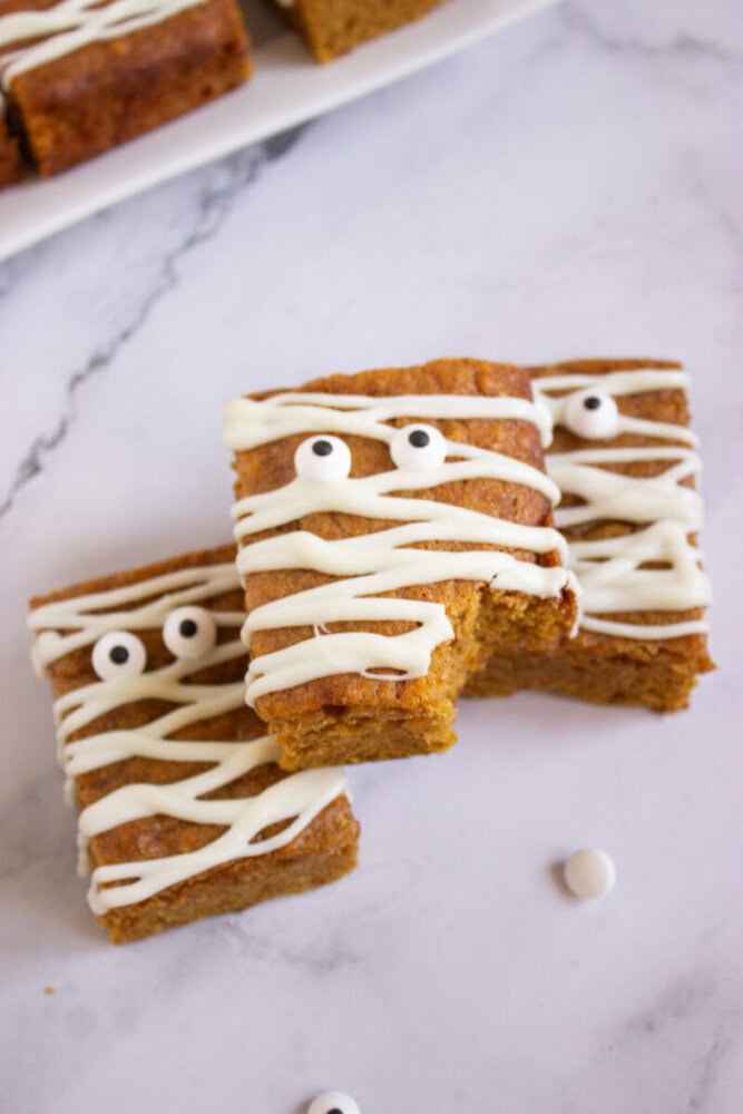 Three pumpkin bars decorated with white icing to resemble mummies, topped with candy eyes, are arranged on a marble surface.