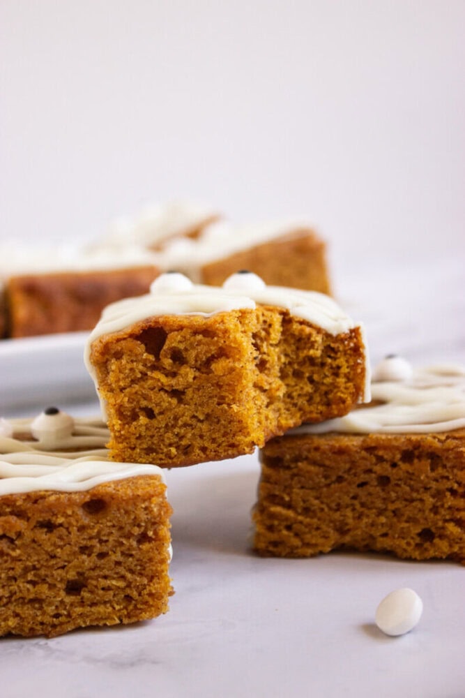 A close-up of a slice of pumpkin cake with white icing on top, held above other pieces on a white surface.
