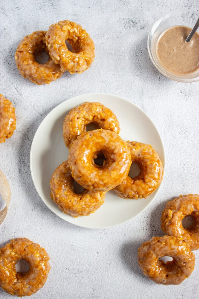 A plate of glazed donuts on a white surface, with extra donuts around the plate and a bowl of glaze with a spoon nearby.