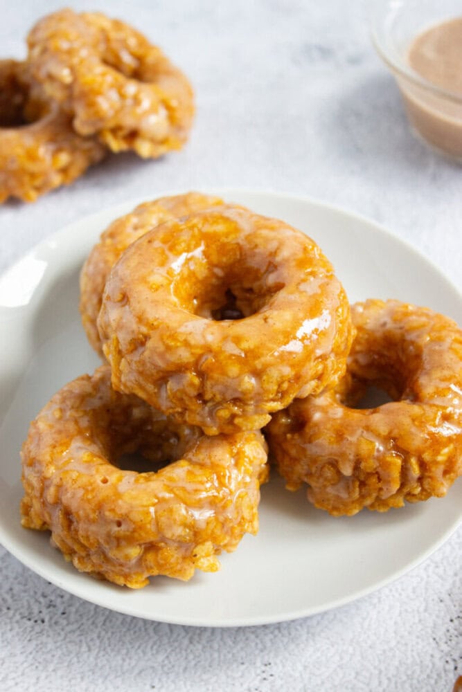 A plate with five glazed, round rice crispy donuts stacked on top of each other, with another donut and a bowl of glaze in the background.