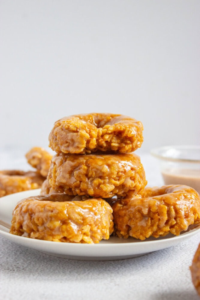 A stack of glazed pumpkin donuts is arranged on a white plate, with more donuts and a small bowl of glaze in the background.