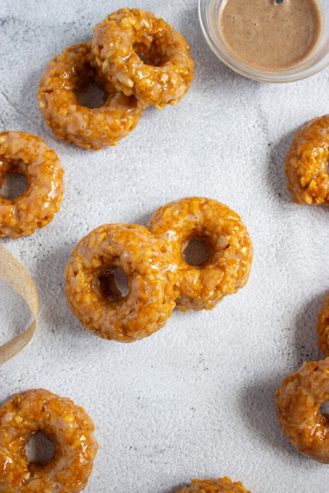 Several glazed donuts are arranged on a light-colored surface, with a small bowl of brown dipping sauce in the top right corner.