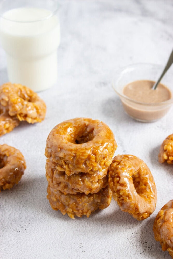 A stack of glazed donuts sits on a light surface, surrounded by more donuts, a glass of milk, and a small bowl of sauce with a spoon.
