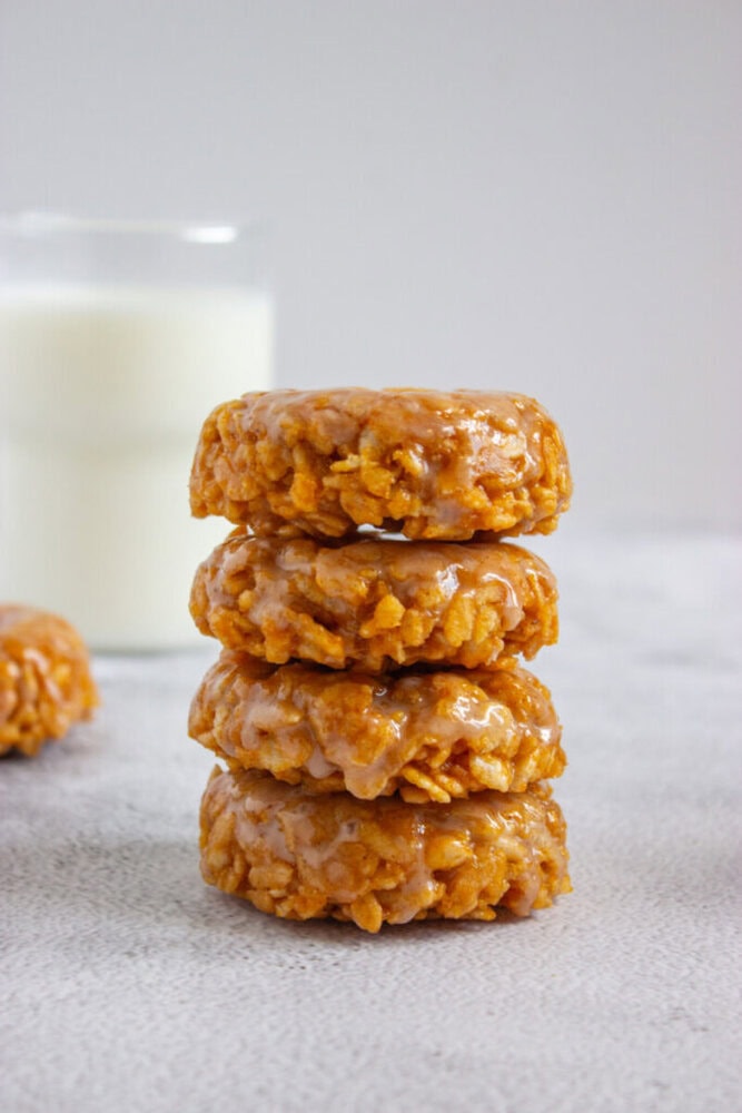 A stack of four glazed oatmeal cookies sits on a light surface, with a glass of milk in the background.