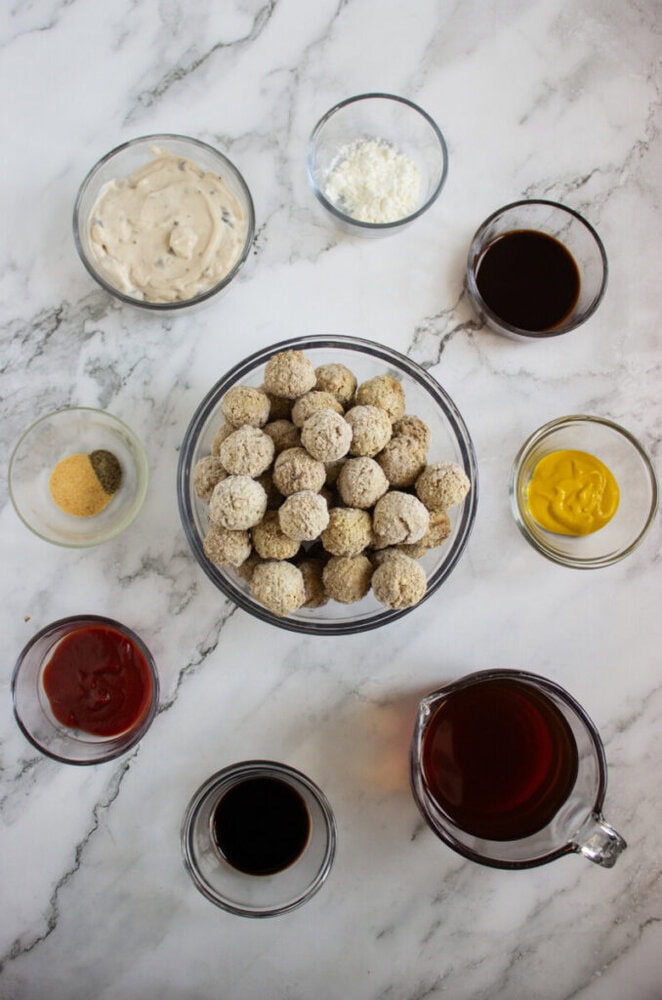 A bowl of uncooked meatballs is surrounded by small bowls of various sauces, seasonings, and ingredients on a marble surface.