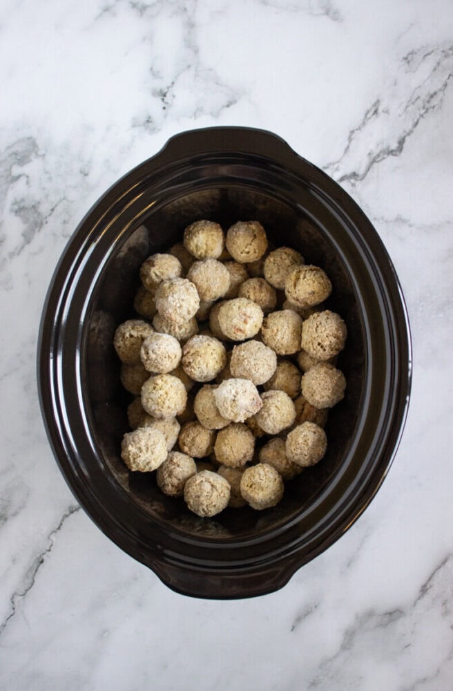 A black slow cooker filled with uncooked meatballs, placed on a white marble surface.