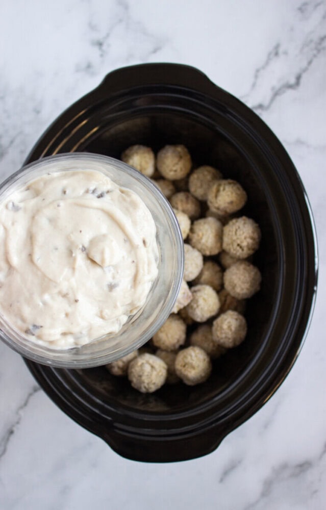 A bowl of creamy sauce is being poured over a layer of frozen meatballs inside a black slow cooker on a marble surface.