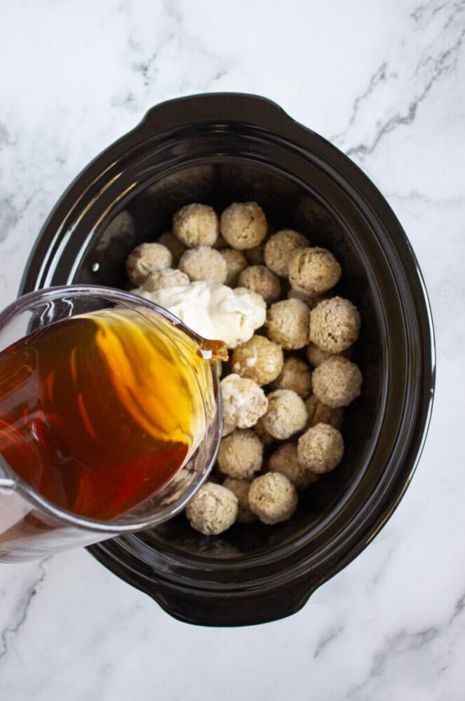 A slow cooker filled with frozen meatballs and cream sauce, with a pitcher of brown liquid being poured over them on a marble countertop.