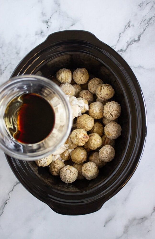 A crockpot filled with frozen meatballs, cream sauce, and a glass measuring cup pouring a dark liquid over them on a marble surface.