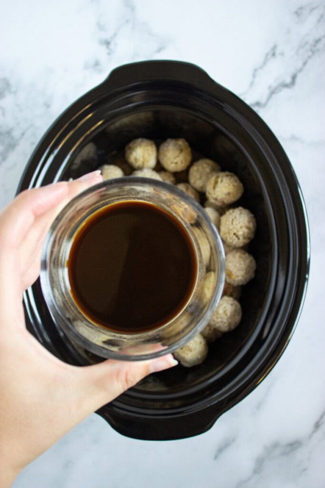A hand holds a bowl of brown sauce above a slow cooker filled with uncooked meatballs on a marble surface.