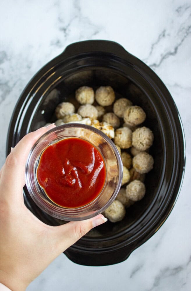 A hand holds a bowl of tomato sauce above a slow cooker filled with frozen meatballs on a marble countertop.