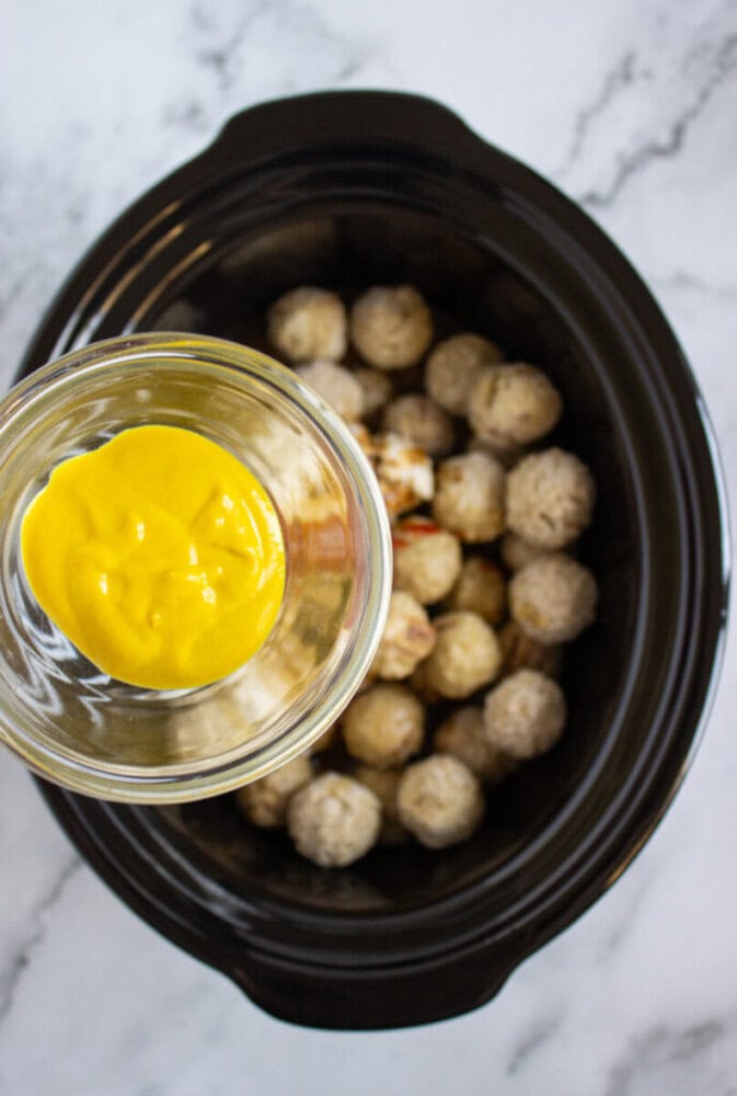 A glass bowl of yellow mustard is held above a slow cooker filled with frozen meatballs on a marble countertop.