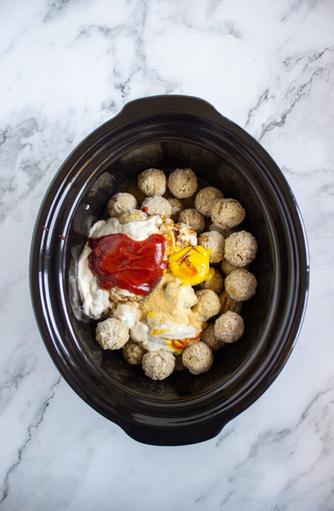 Frozen meatballs in a slow cooker topped with ketchup, mayonnaise, mustard, and seasoning, ready to be mixed and cooked.