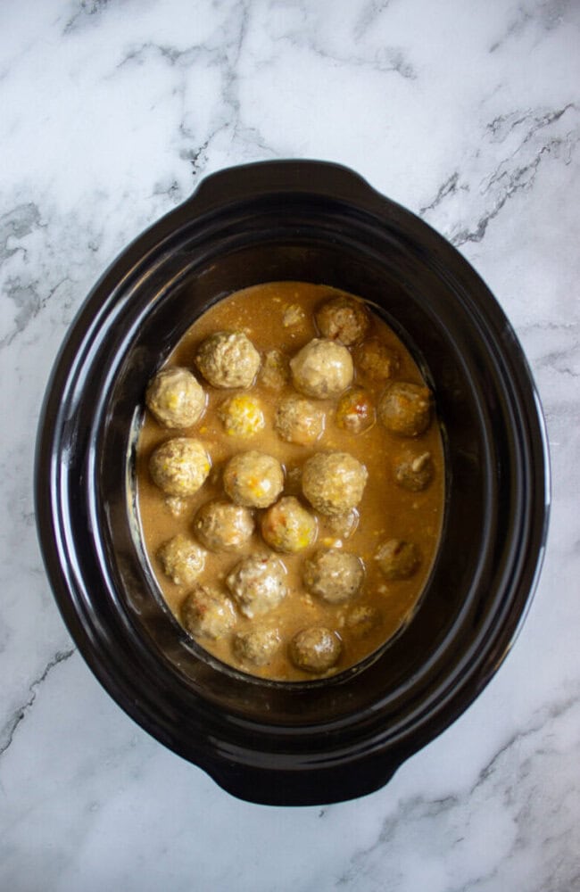 Meatballs in a creamy brown sauce inside a black slow cooker, placed on a white marble surface.
