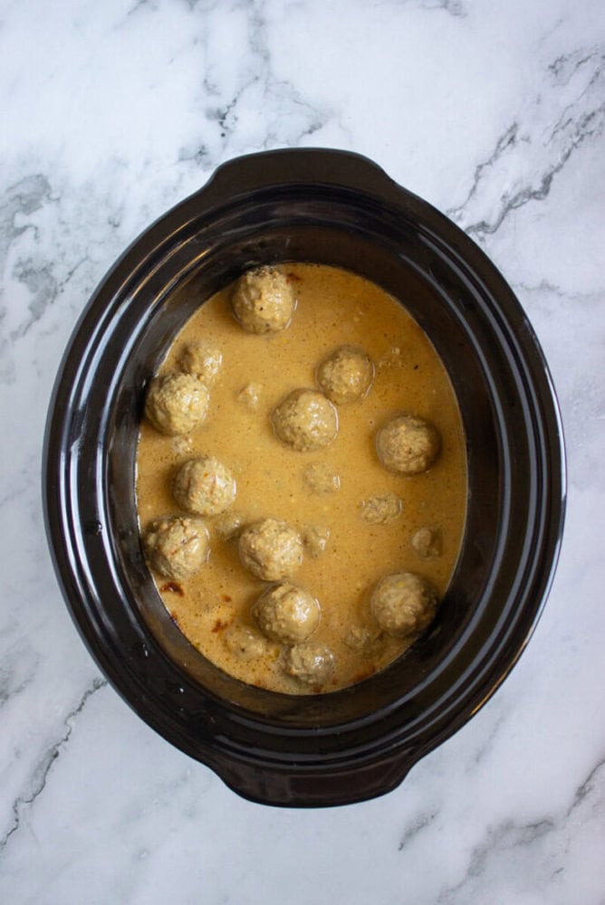 A black slow cooker containing meatballs in a creamy beige sauce sits on a white marble surface.