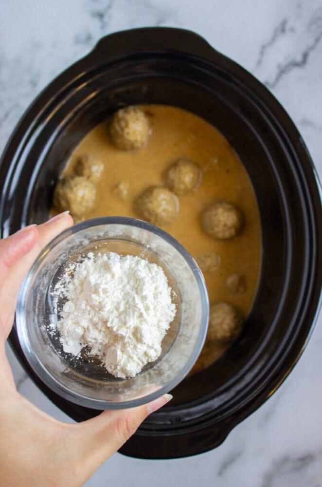 A hand holds a small glass bowl of white cornstarch above a slow cooker containing meatballs in a brown sauce.