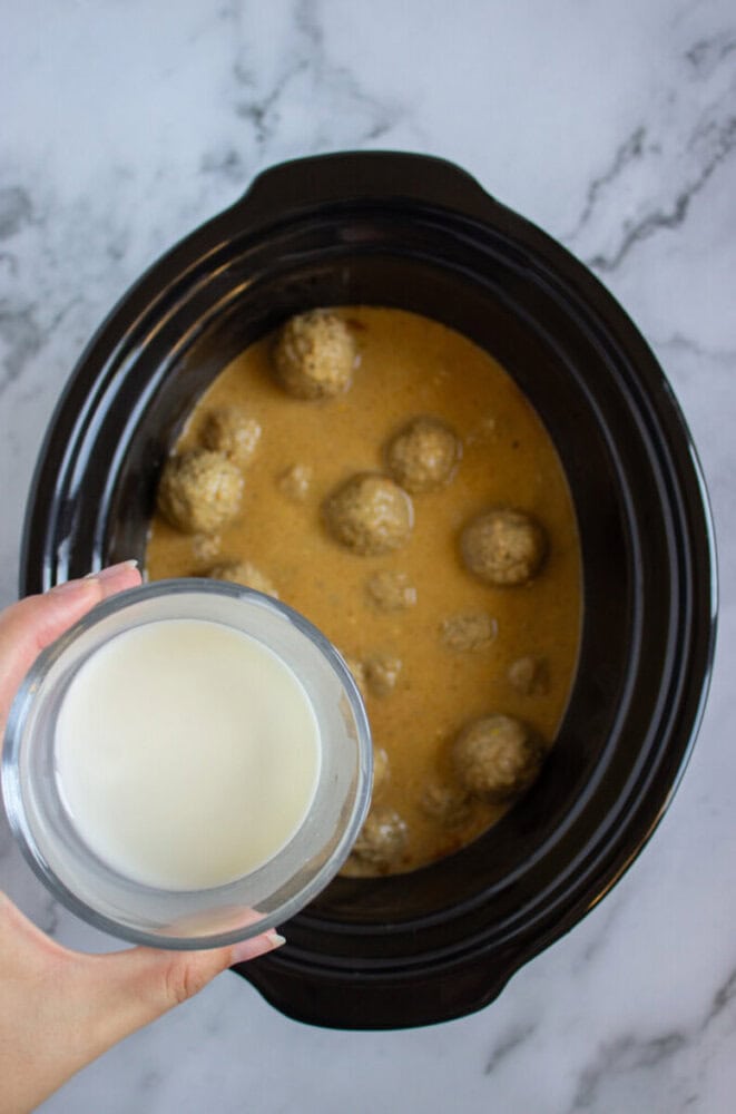 A hand holds a bowl of milk above a slow cooker containing meatballs in a creamy sauce on a marble surface.