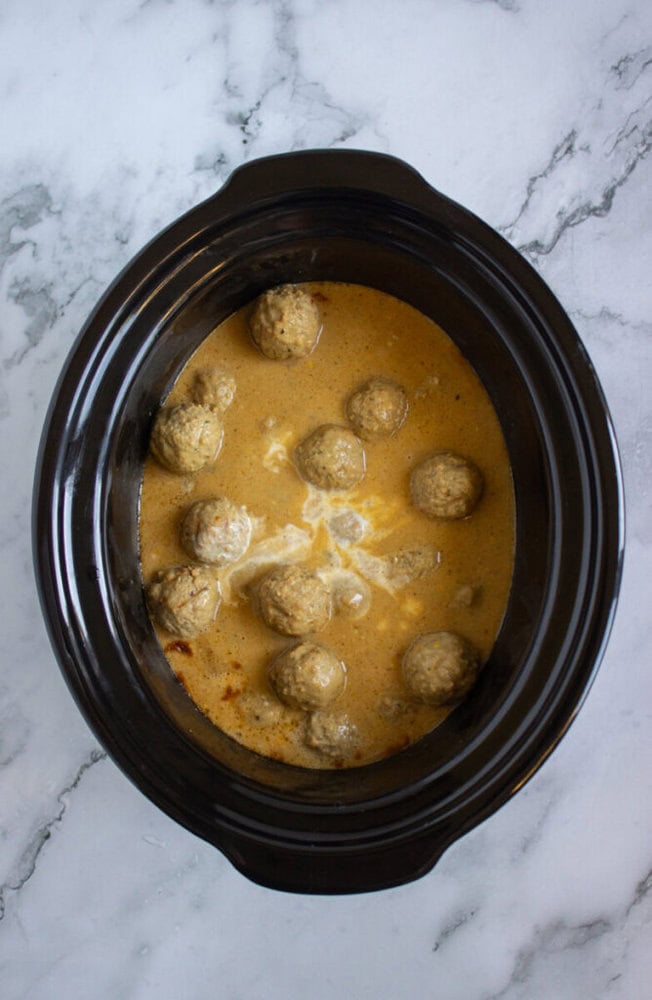 Meatballs in a creamy sauce inside a black slow cooker, placed on a white marble surface.
