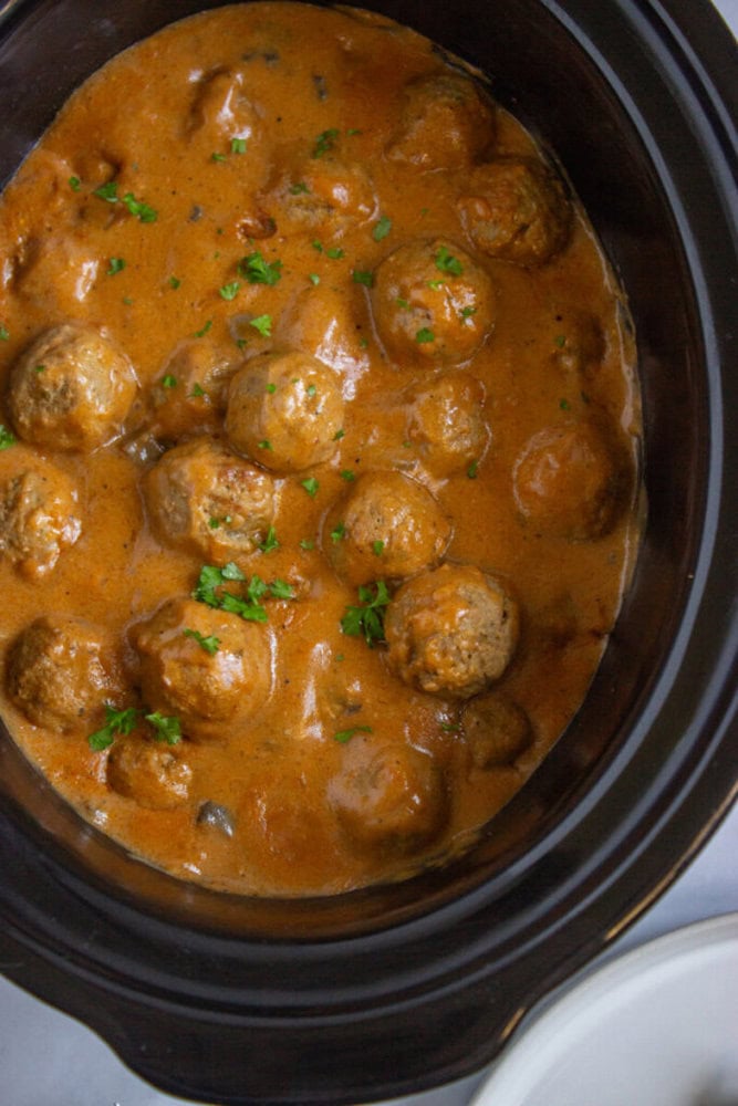 Meatballs in a creamy brown sauce garnished with chopped parsley, served in a black slow cooker.