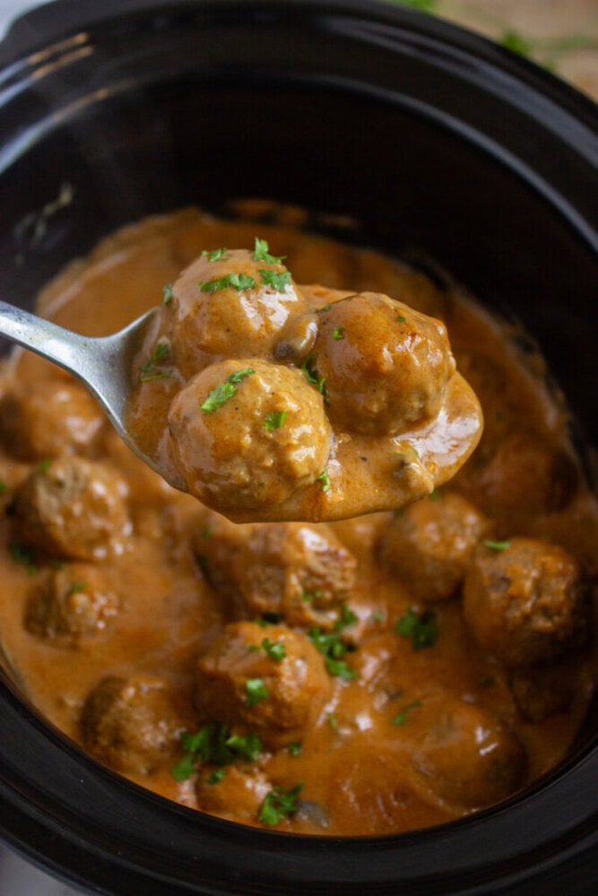 A spoonful of meatballs in creamy sauce is held over a slow cooker filled with more meatballs, garnished with chopped herbs.